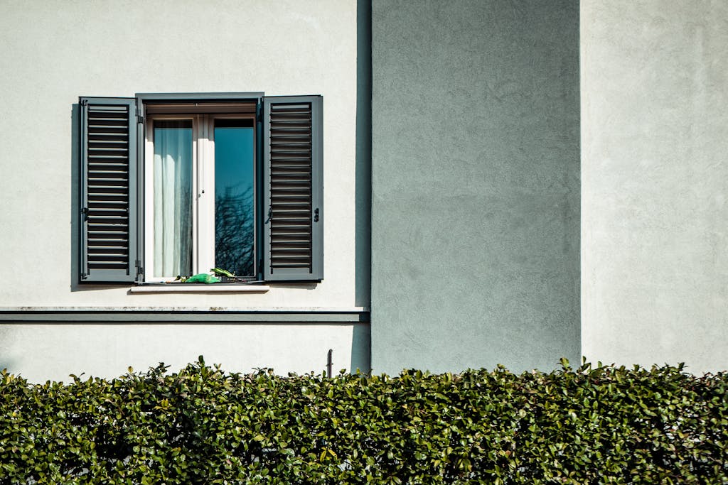 A minimalist exterior featuring a window with open shutters against a white wall with green hedges.